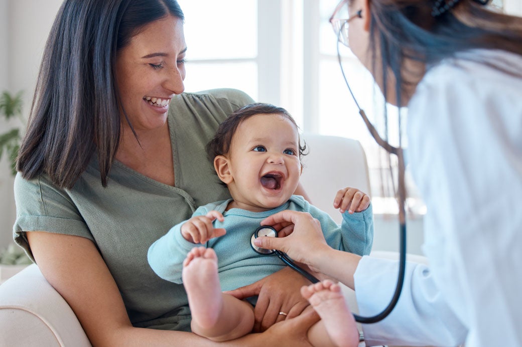 Doctor with stethoscope examining smiling baby who is held by smiling mom