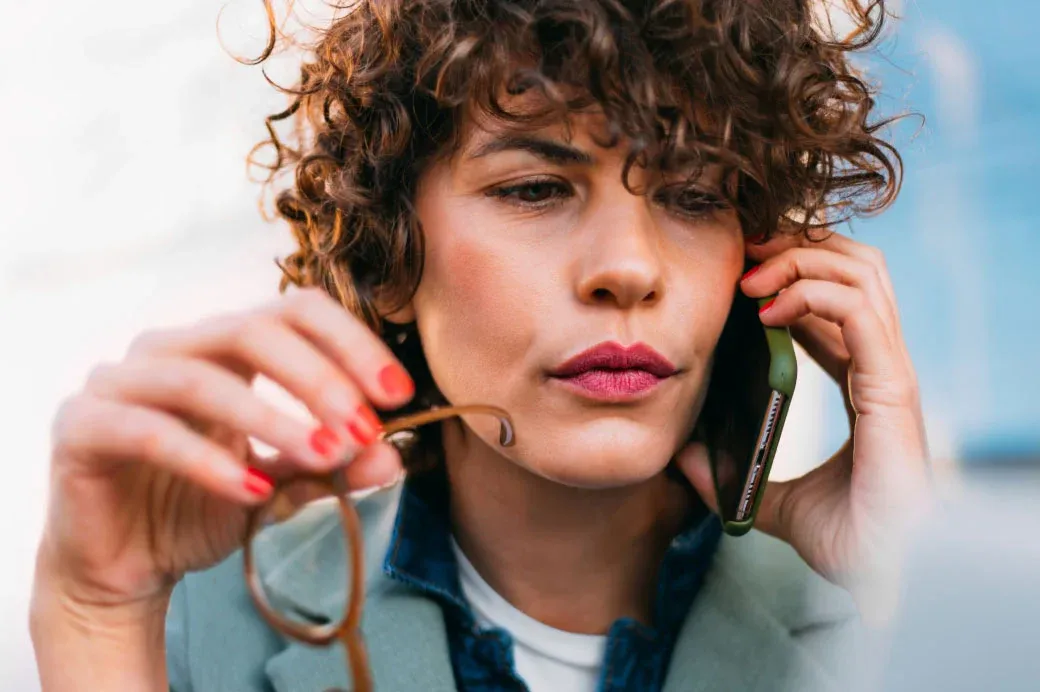 Woman with curly hair holding eyeglasses while talking on a mobile phone