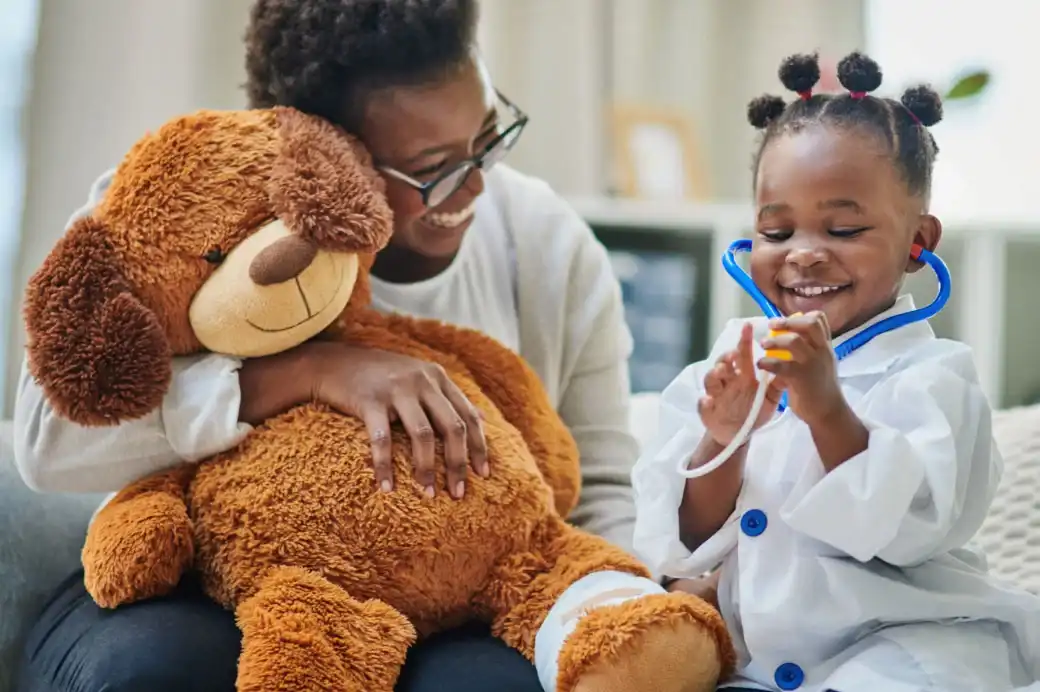 Woman holding a teddy bear smiling at a child pretending to be a healthcare worker.