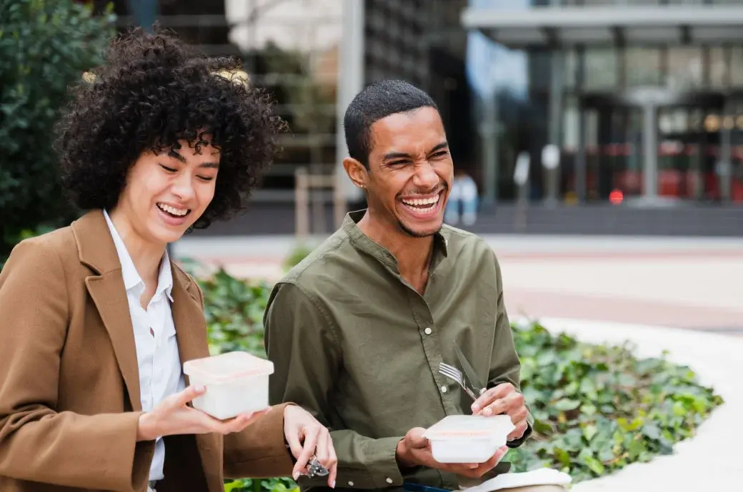 A woman and man sitting next to one another in an outdoor courtyard laughing and preparing to eat food from containers